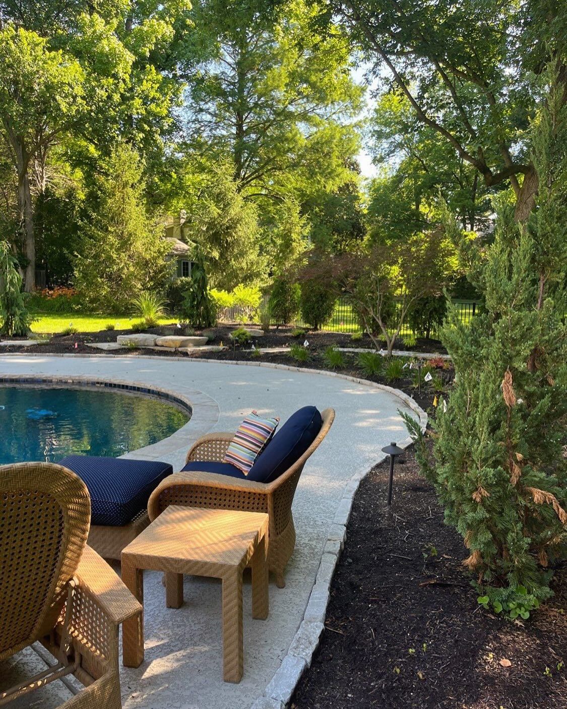 Wicker furniture with blue cushions beside a curved pool. A garden path and trees surround the pool.