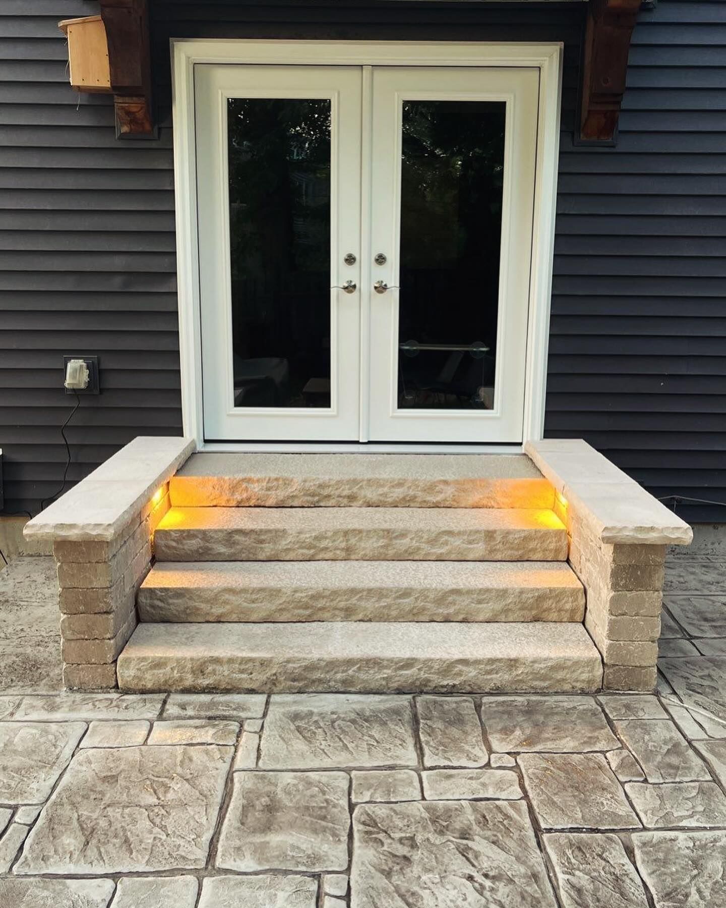 Stone steps with built-in lights lead to white double doors. The doors are framed by dark siding, 