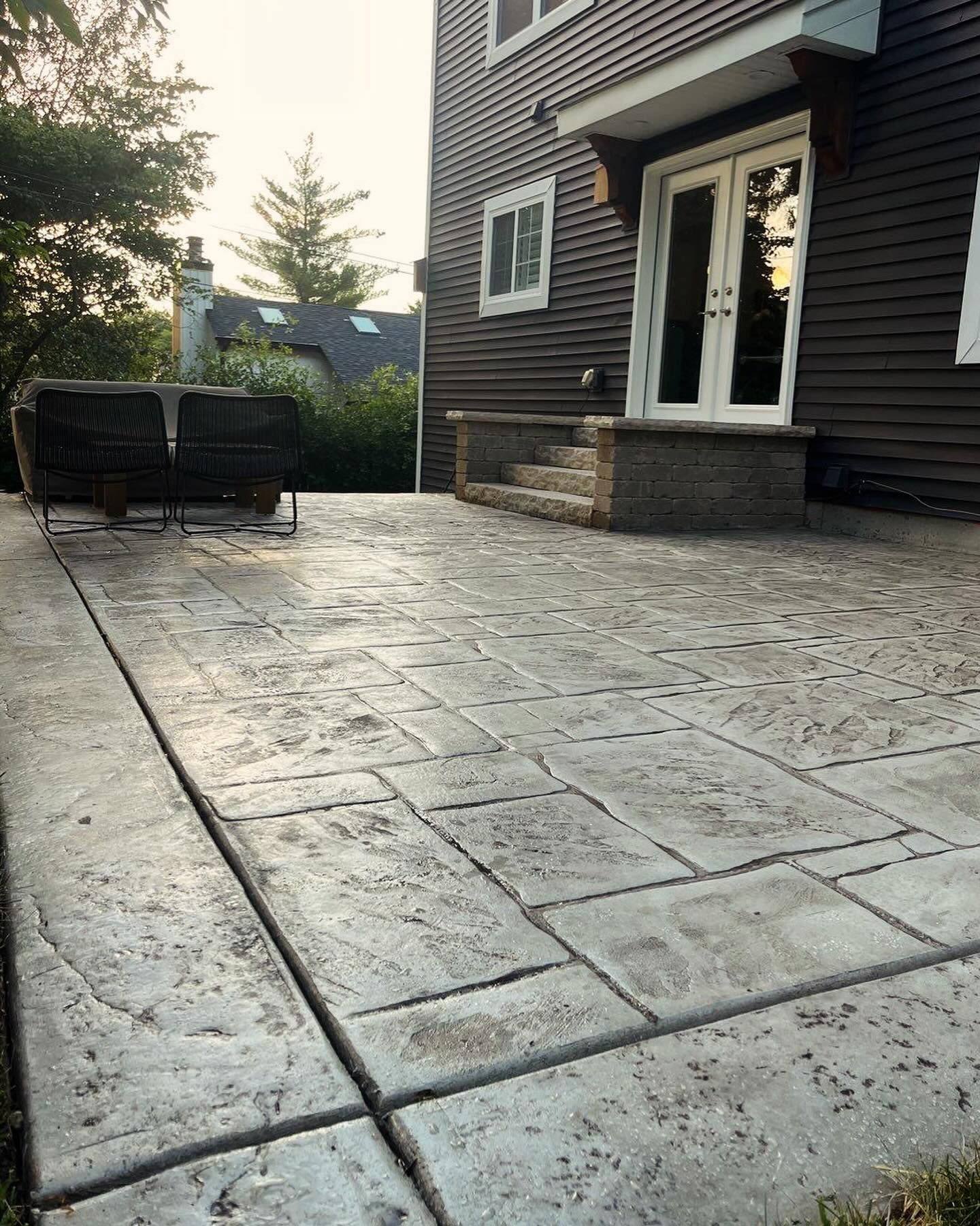 A stamped concrete patio in front of a dark-sided house with a wicker couch and double doors.