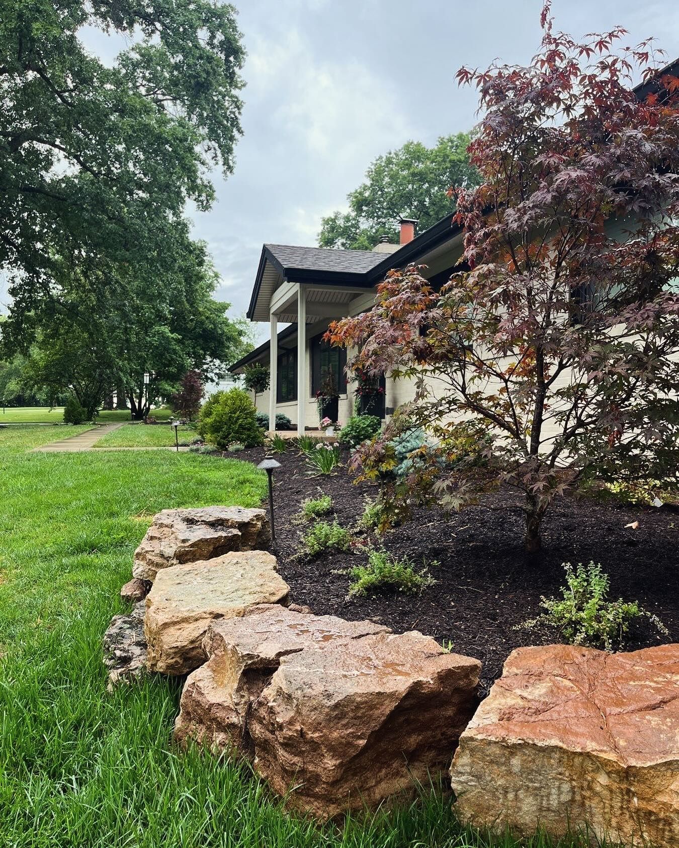A house with a porch is next to a rock garden with dark mulch and colorful plants, a grassy lawn, and trees.