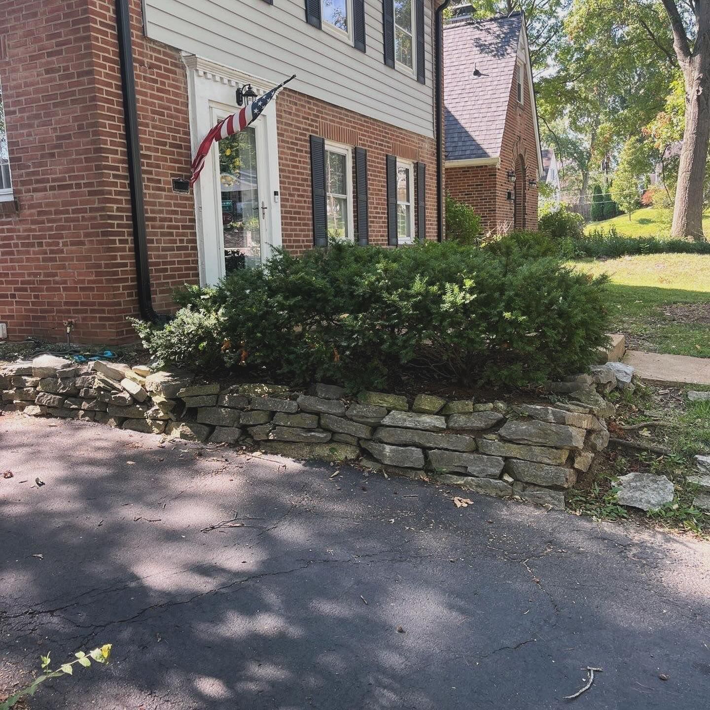 Brick house with a stone retaining wall and a large green bush in front. An American flag hangs from a window.