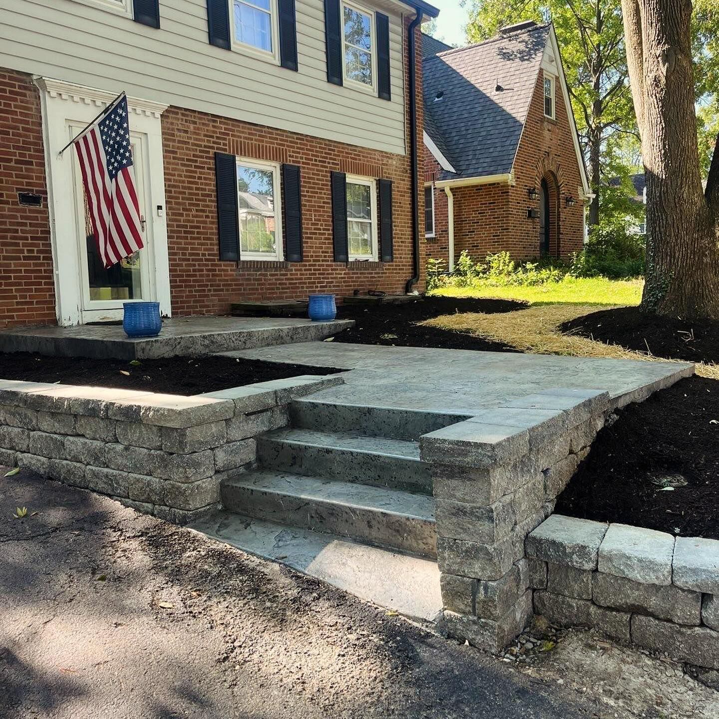 A brick house with a stone patio and steps leading to the front door, an American flag hangs on the door.