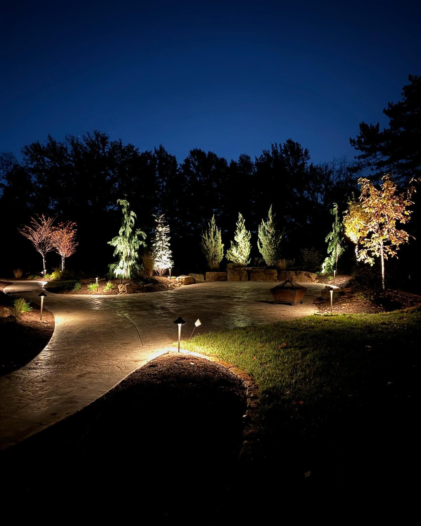 Nighttime outdoor scene with garden lighting illuminating a winding path, trees, and shrubs against a dark sky.
