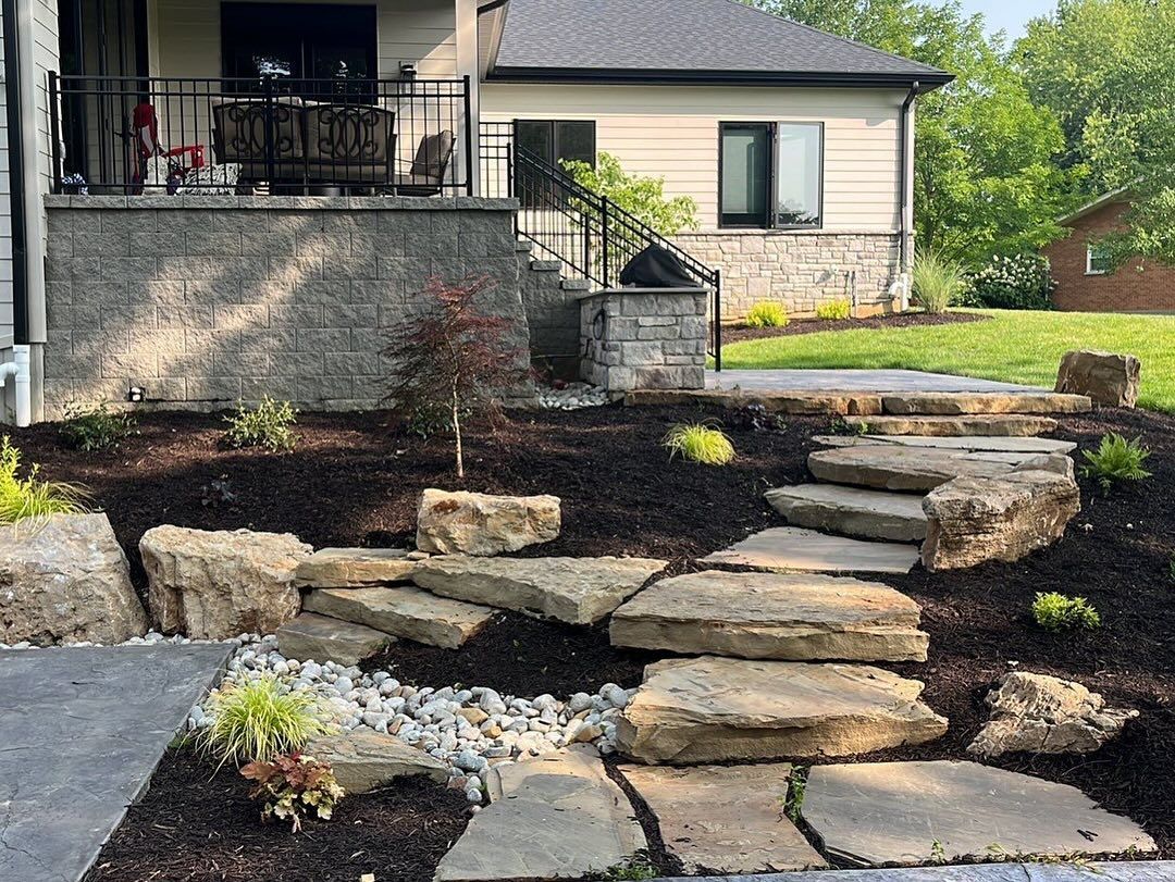 Stone steps and landscaping leading up to a house with a patio. Dark mulch, small plants, and a mature tree frame the path.