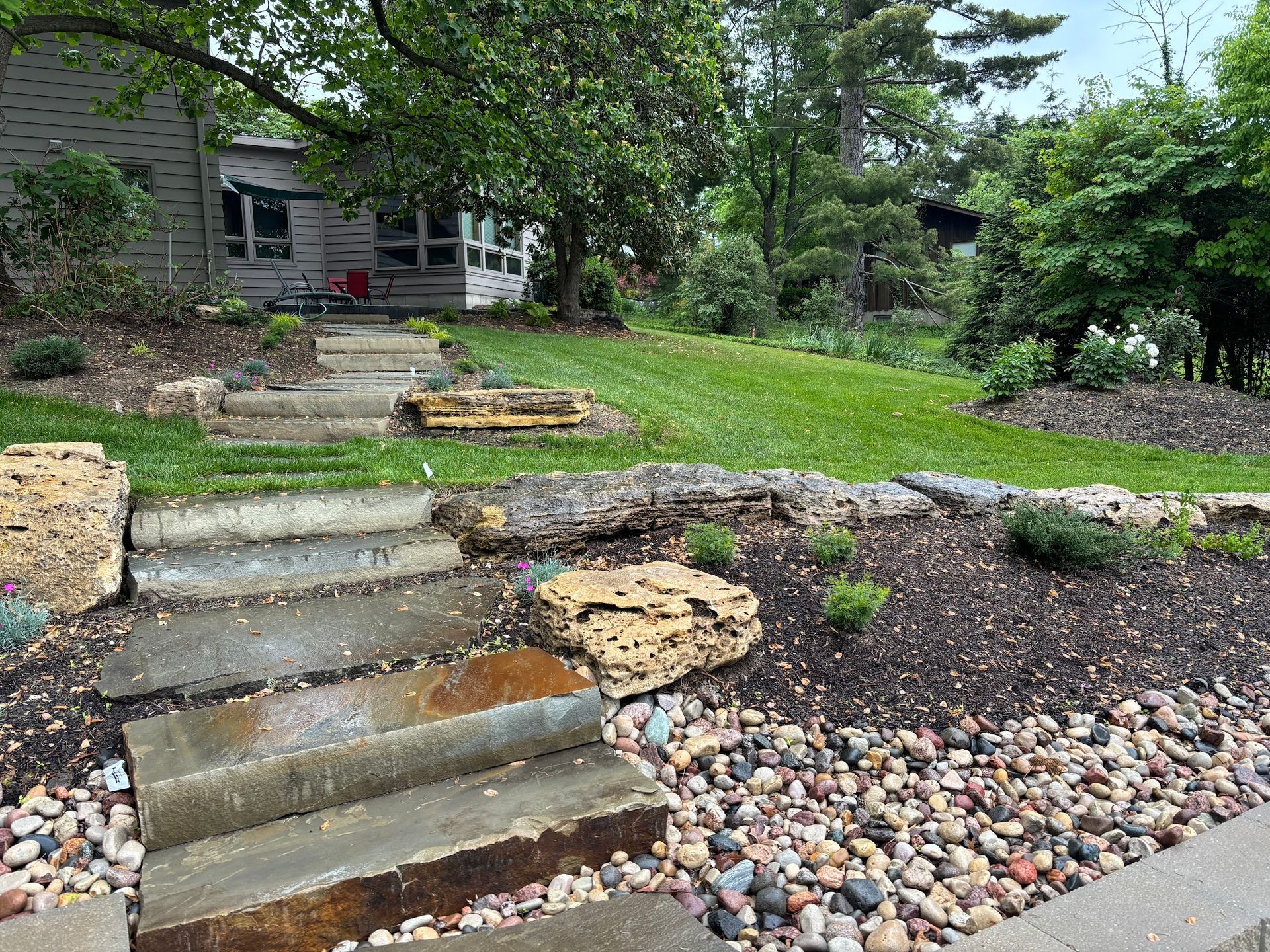 Stone steps leading up to a house with a landscaped yard. The steps are wet with some large rocks and gravel in front.