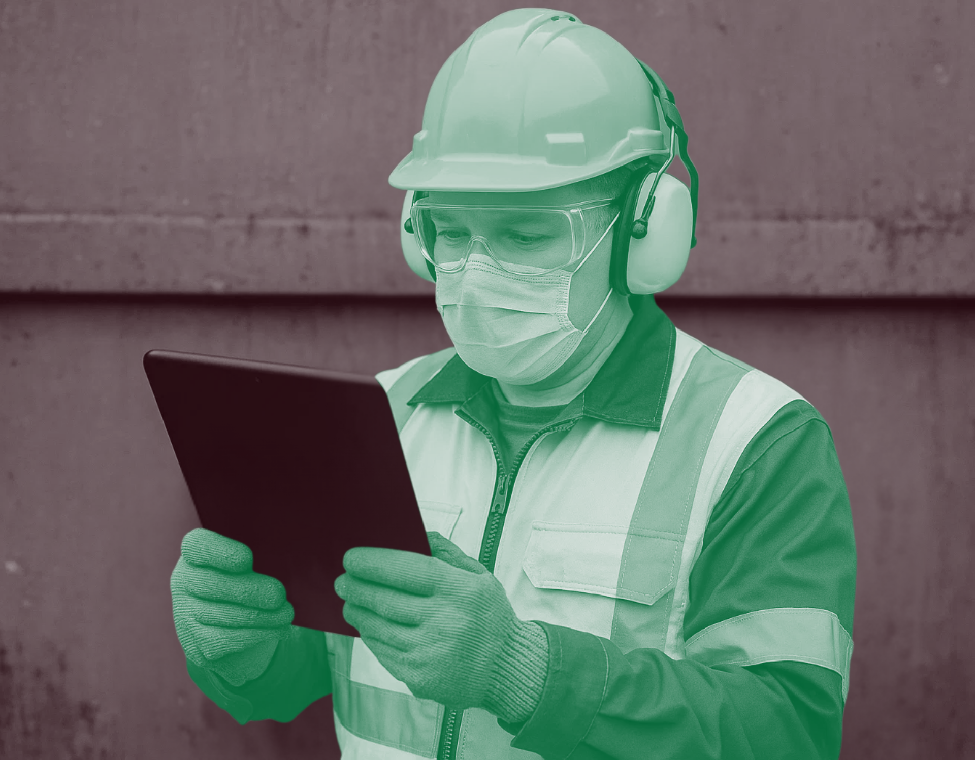 A man wearing a hard hat , ear muffs and a mask is looking at a tablet.