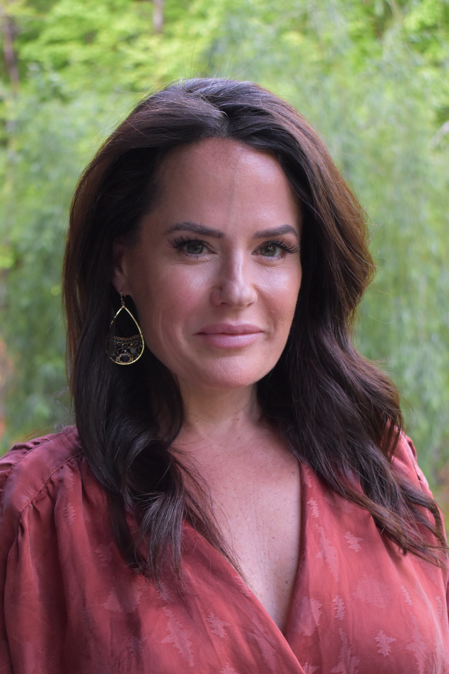 Woman with dark hair wearing a rust-colored dress and gold earrings, posing outdoors.