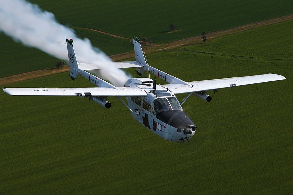 A small plane is flying over a field with smoke coming out of it