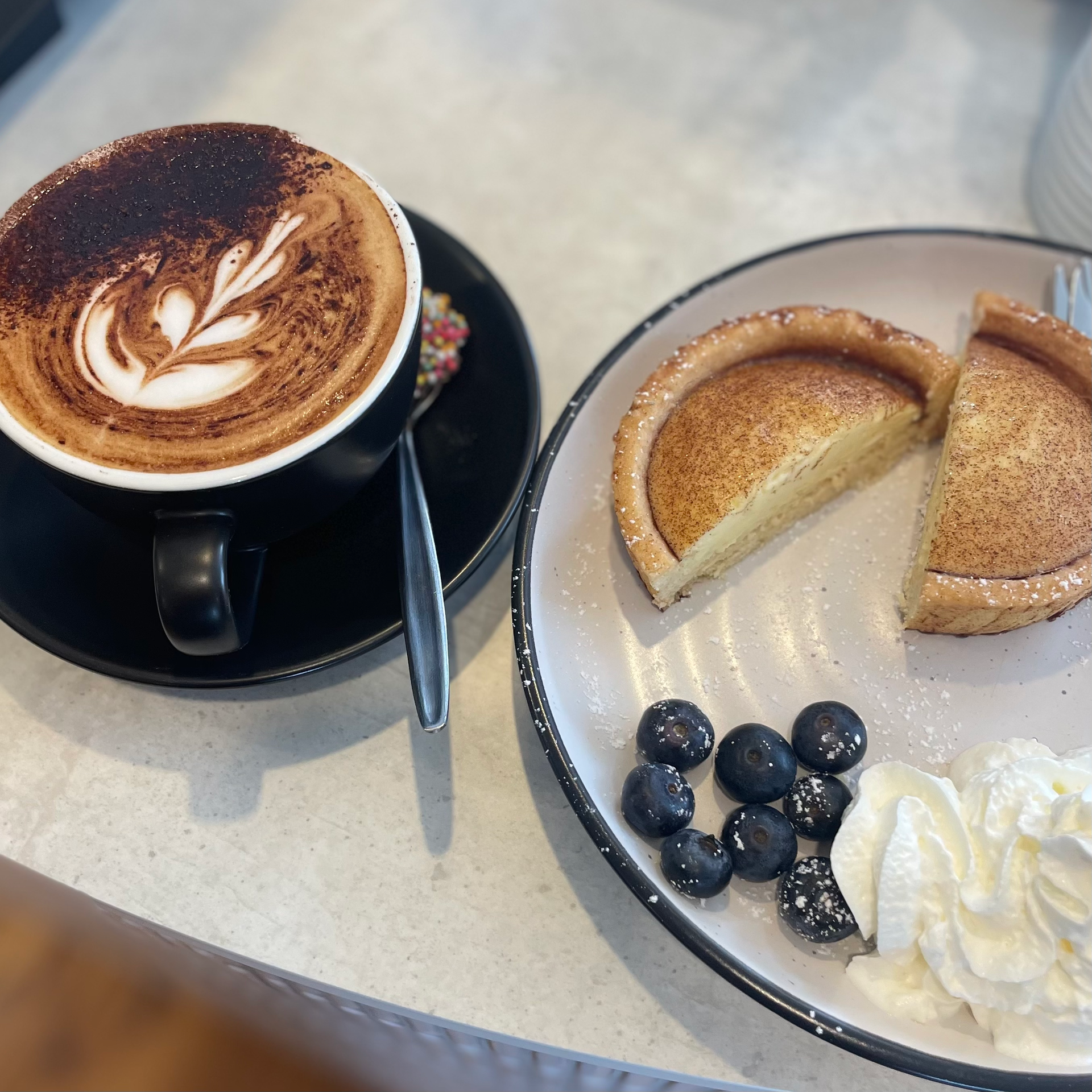 A cup of coffee next to a plate of food with whipped cream and blueberries