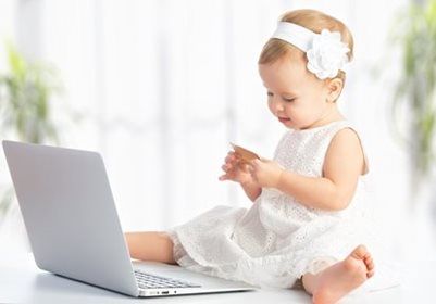 A child wearing a white dress and headband sits on a floor holding a credit card while looking at an open laptop.