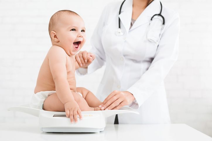 A doctor in a white coat weighs a happy, sitting infant on a clinical scale.