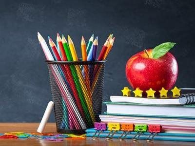 A wire cup filled with colorful pencils sits next to books topped with a red apple, featuring small star and face clips.