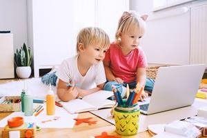 Two children lying on the floor, using a laptop together surrounded by art supplies and books.