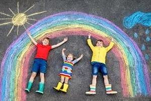 Three children lie on the ground under a large rainbow drawn in chalk, next to a sun and a rain cloud.