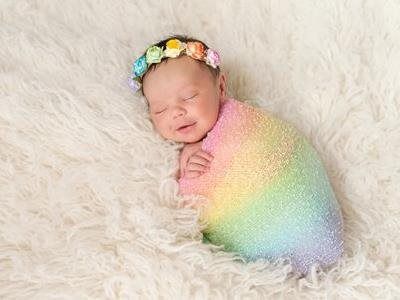 A newborn wrapped in a rainbow-colored blanket, wearing a floral crown, sleeping on a plush white faux fur surface.