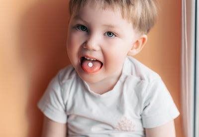 A toddler in a white shirt with a small, round white pill on their tongue, set against a solid orange background.