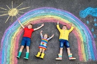 Three children in rain boots pose under a chalk rainbow, sun, and rain clouds on pavement.