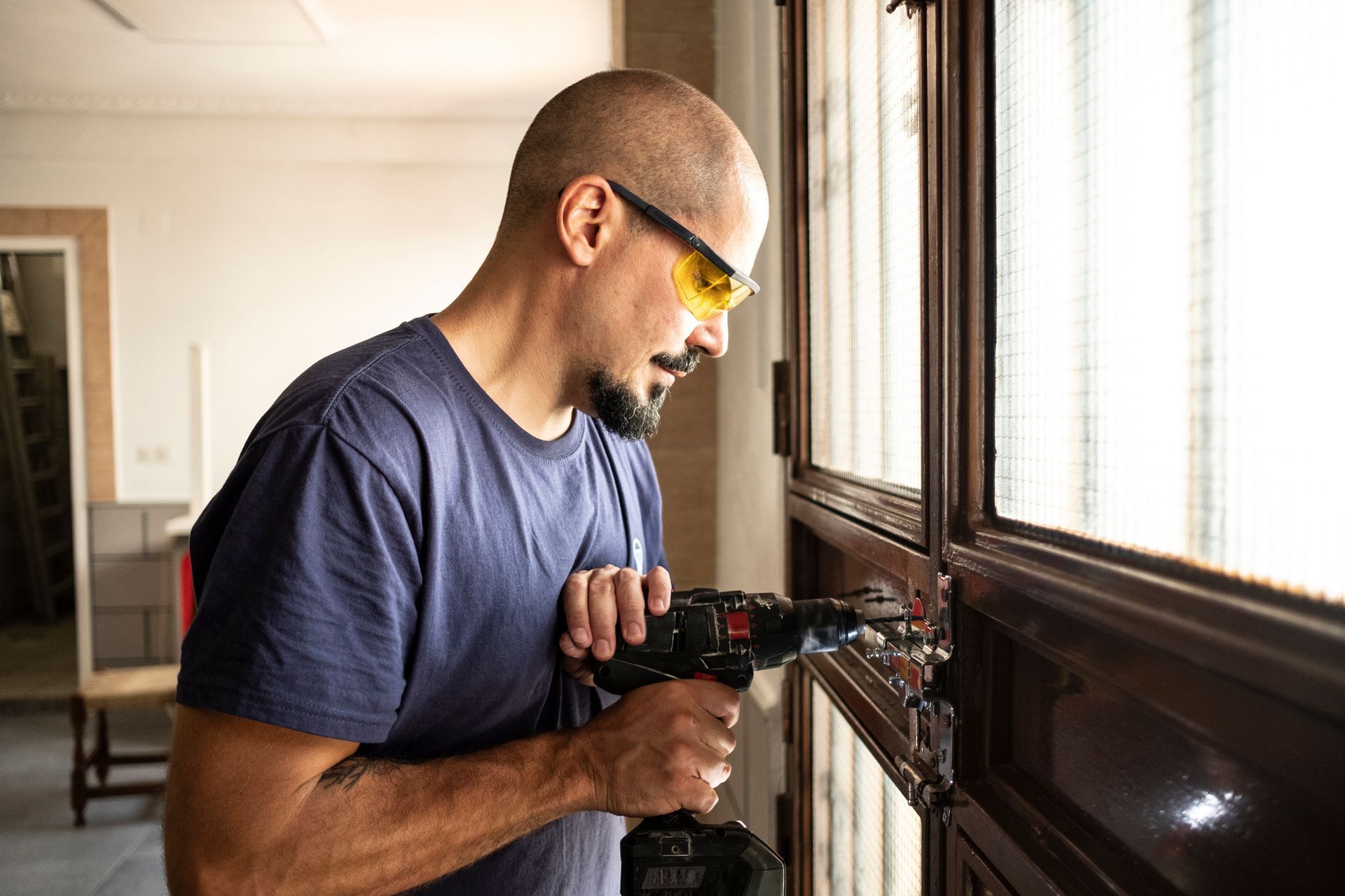 An emergency locksmith wearing protecting eyewear is changing a lock at home with a battery drill.