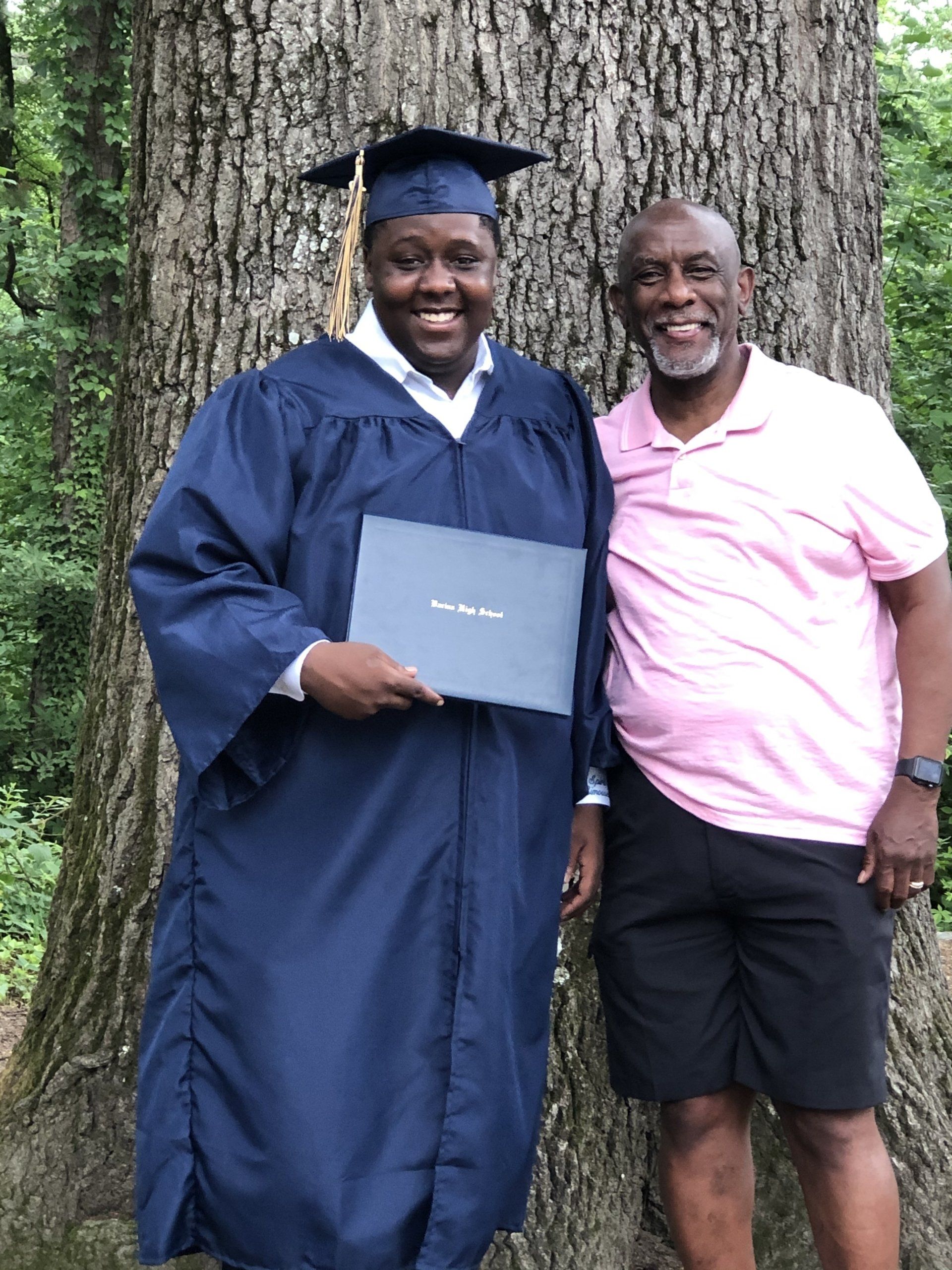 A man in a graduation cap and gown stands next to a man in a pink shirt