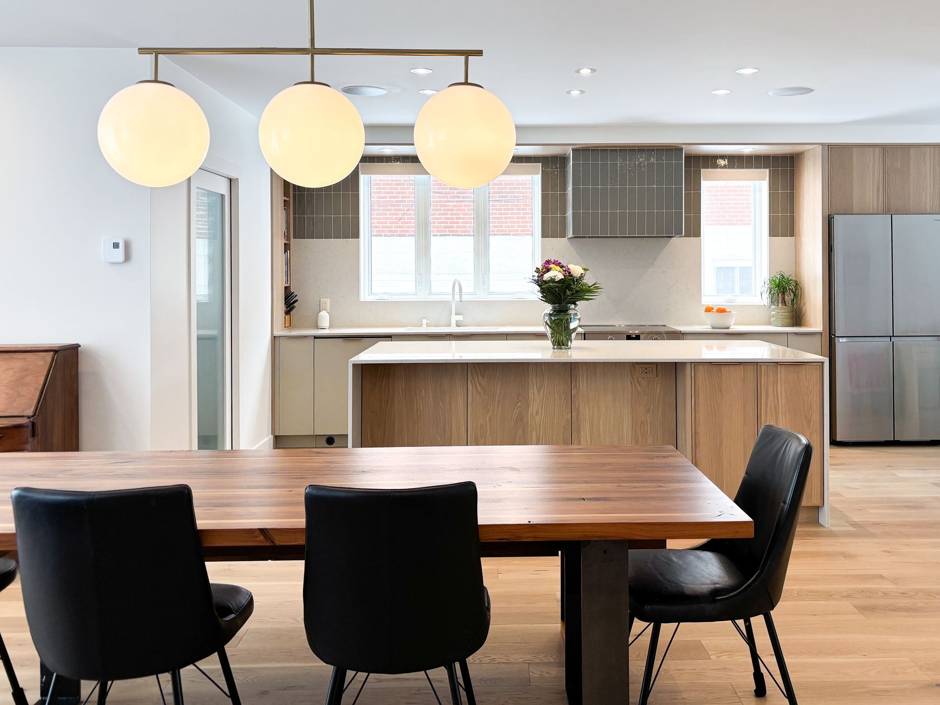 Salle à manger avec table en bois, chaises noires et cuisine moderne avec luminaires suspendus.