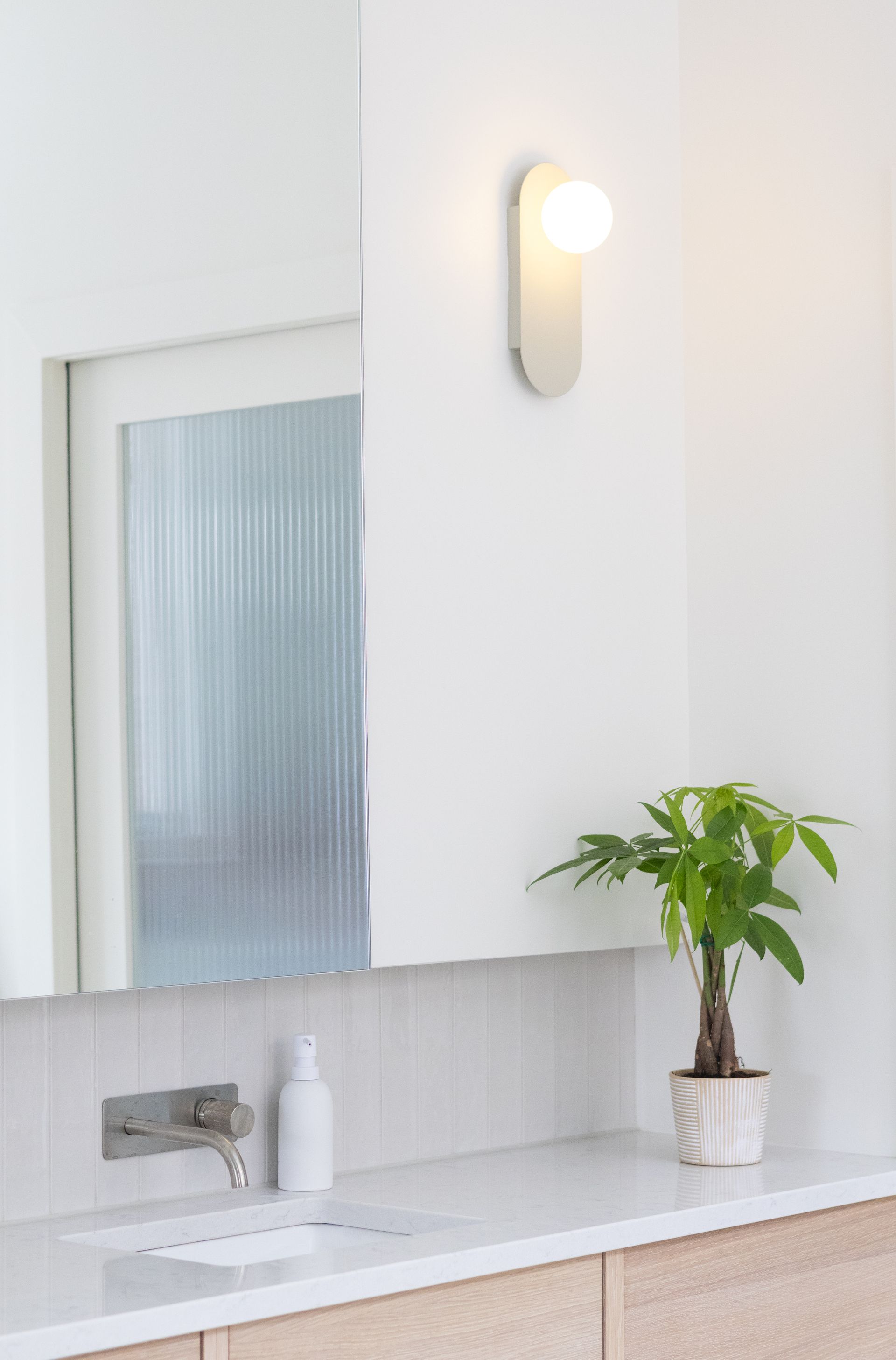 Salle de bain avec plan de travail blanc, miroir et applique murale. Une plante est posée sur le plan de travail.