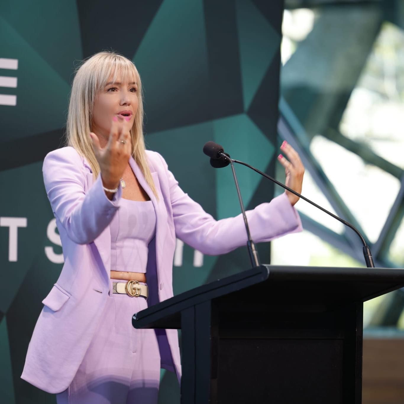 Woman in lavender suit speaks at a podium. She gestures with hands, background is a geometric design.