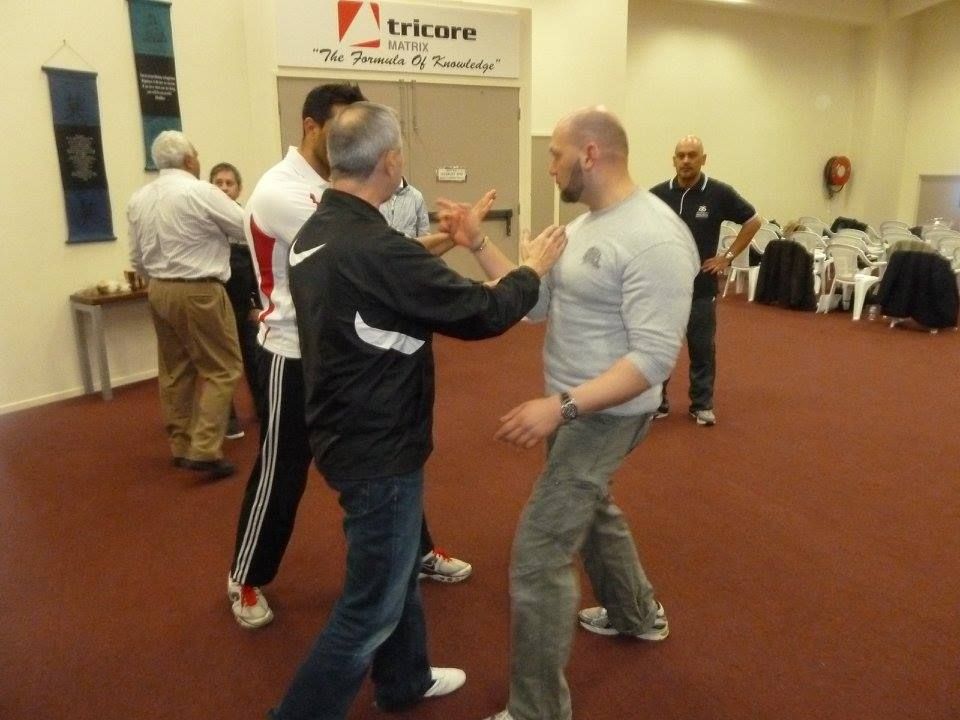 People practicing martial arts in a room with others observing. Two are in contact, demonstrating a move.