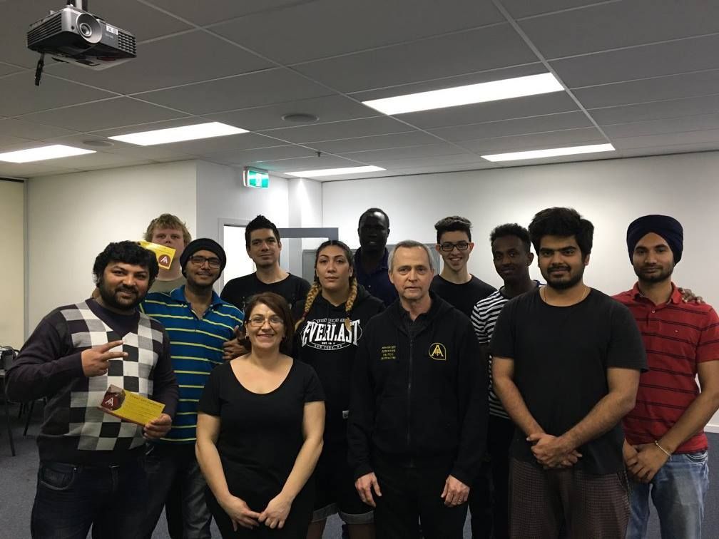 Group of people smiling, posing in a classroom. Most are looking at the camera. Fluorescent lighting, projector visible.