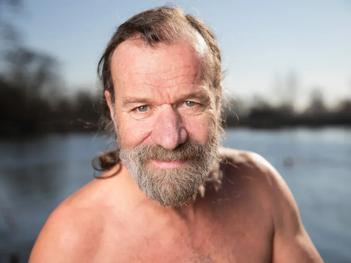 Man with beard stands shirtless near water, smiling.