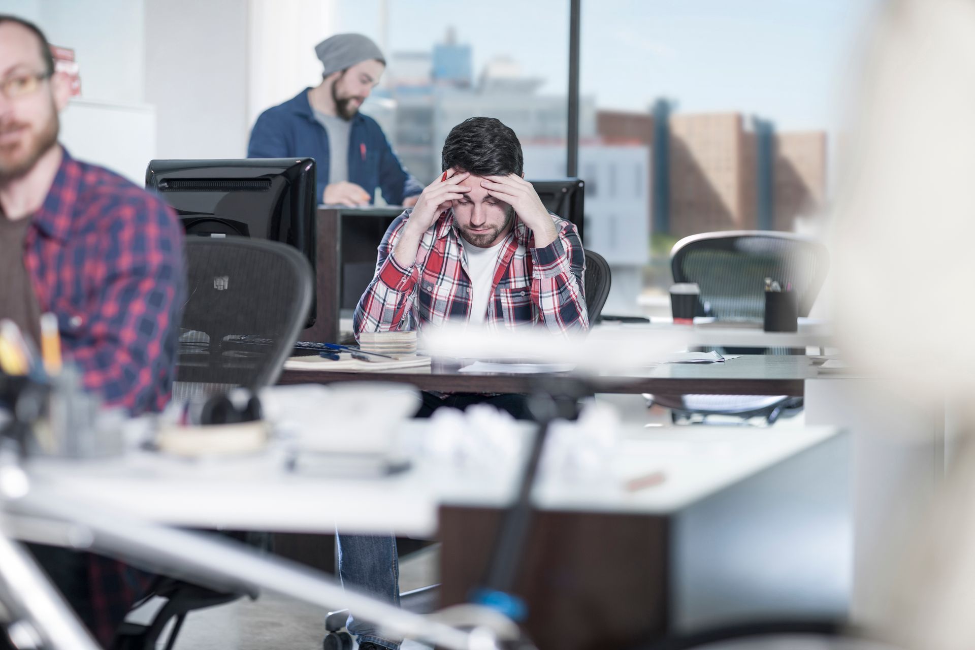 In a bright office, a person in a plaid shirt sits at a desk with their head in their hands, appearing stressed.