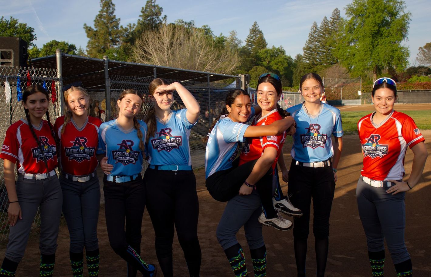 Eight softball players in blue and red jerseys pose outdoors on a field after a game.