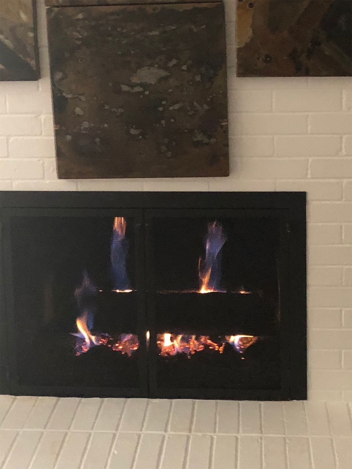 Gas fireplace with blue and orange flames, inside a black frame. White tile surround, art above.