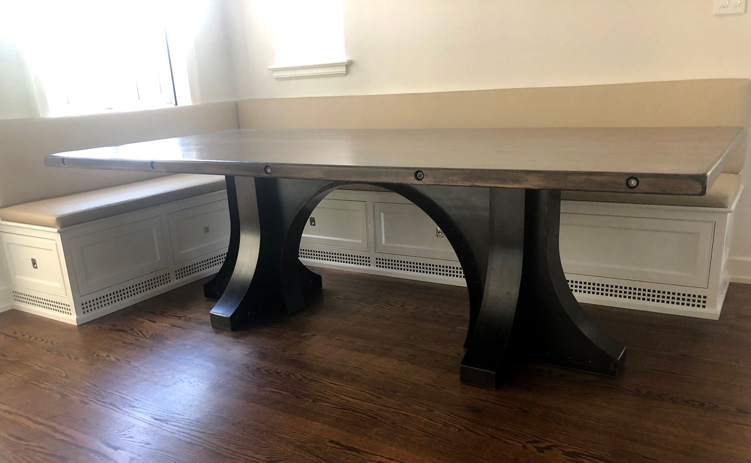 Dining table with dark legs and a light-colored top, placed in front of a built-in bench.