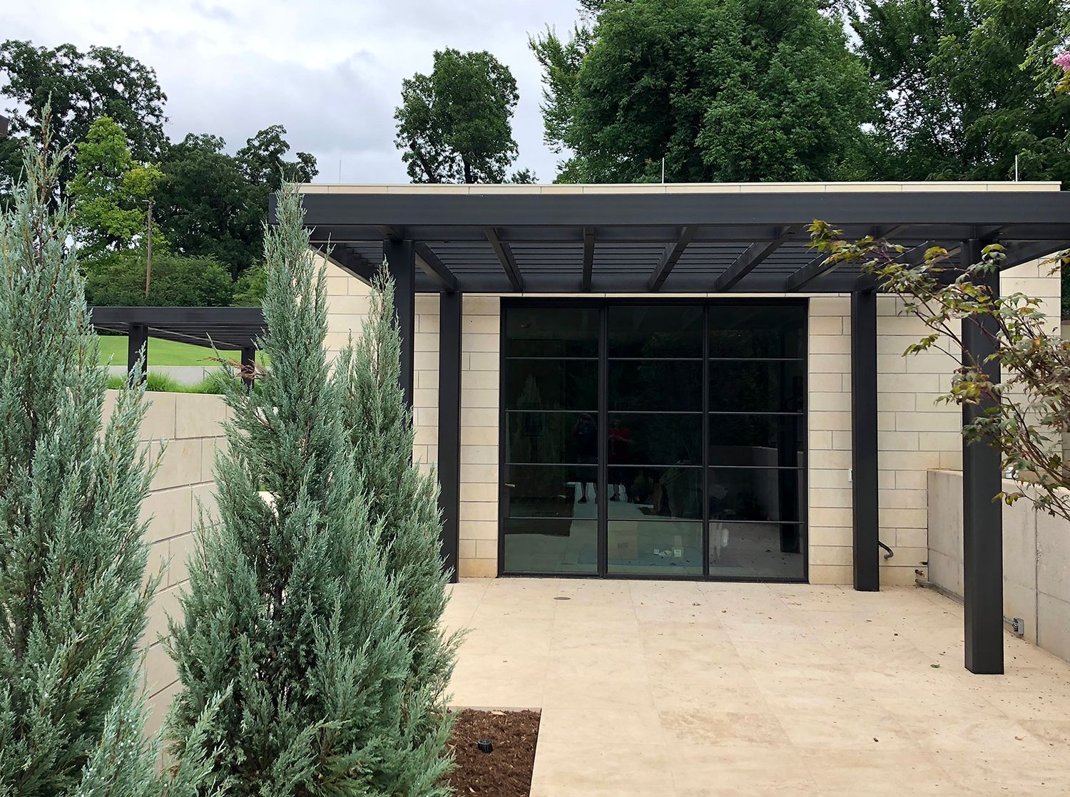 Exterior of a modern building with large windows, covered by a black pergola. Blue-green trees are in the foreground.