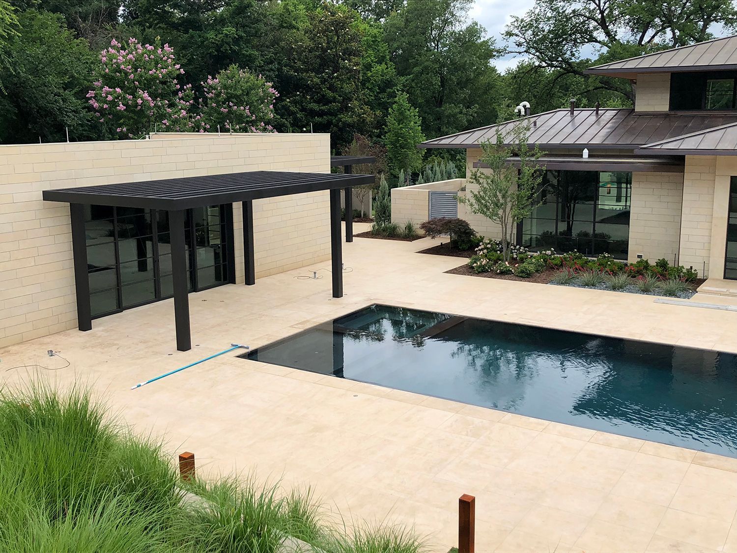 Poolside view: Black pergola, rectangular pool, beige stone patio, modern home, lush greenery.