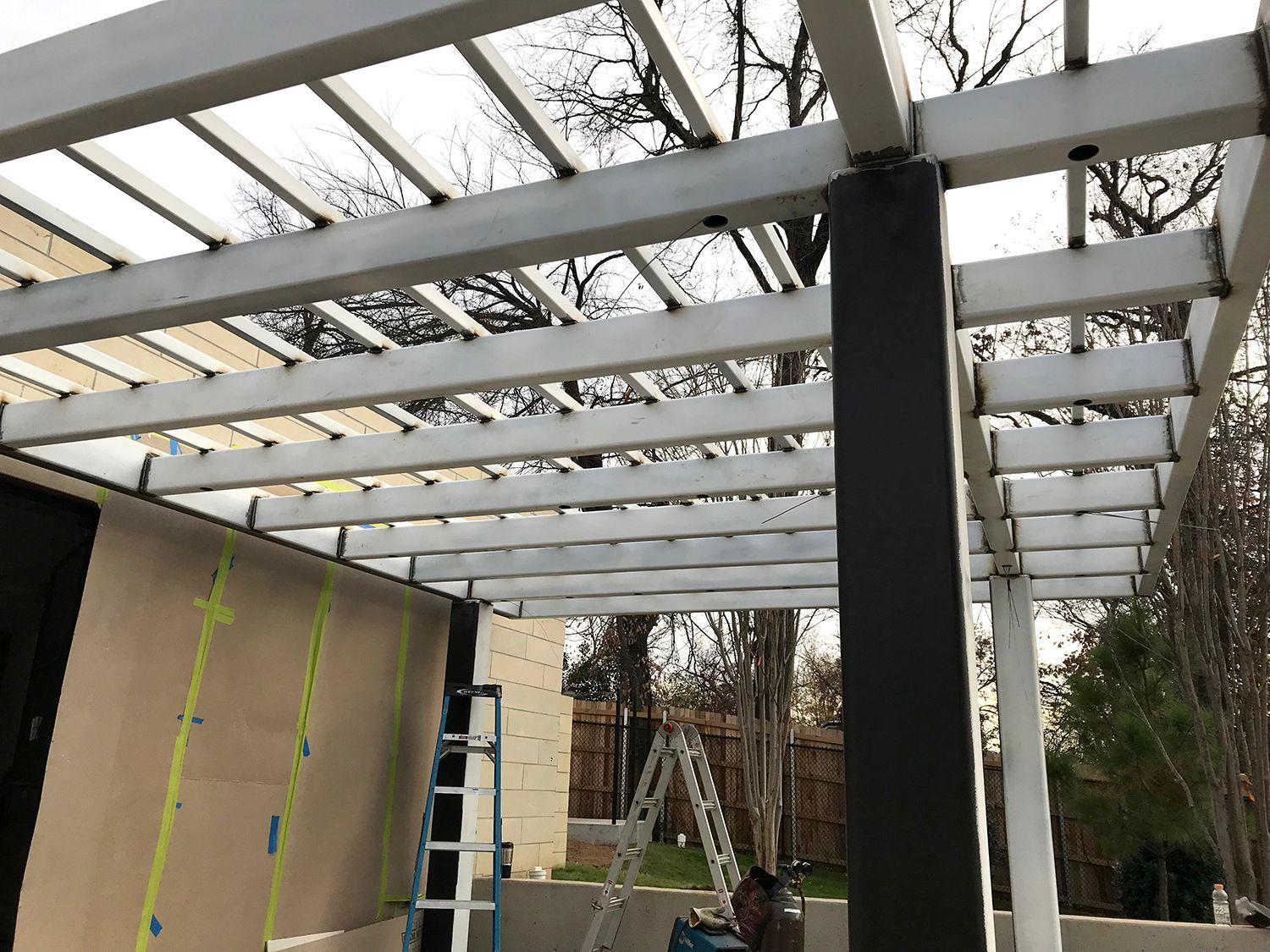 Metal pergola attached to a building, with horizontal beams and a black support post.