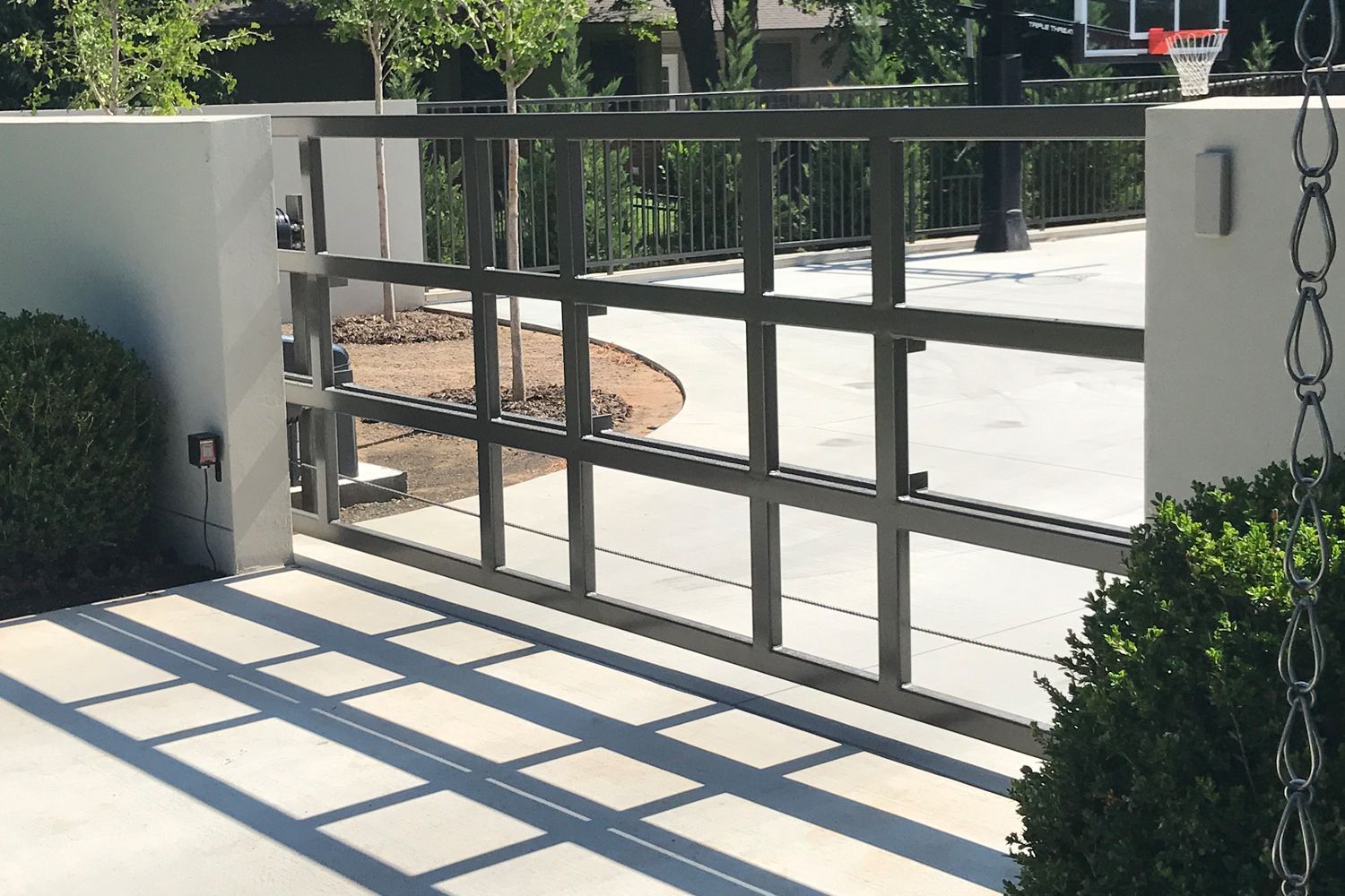 Gray metal gate with square design, between concrete pillars, opening onto a driveway.