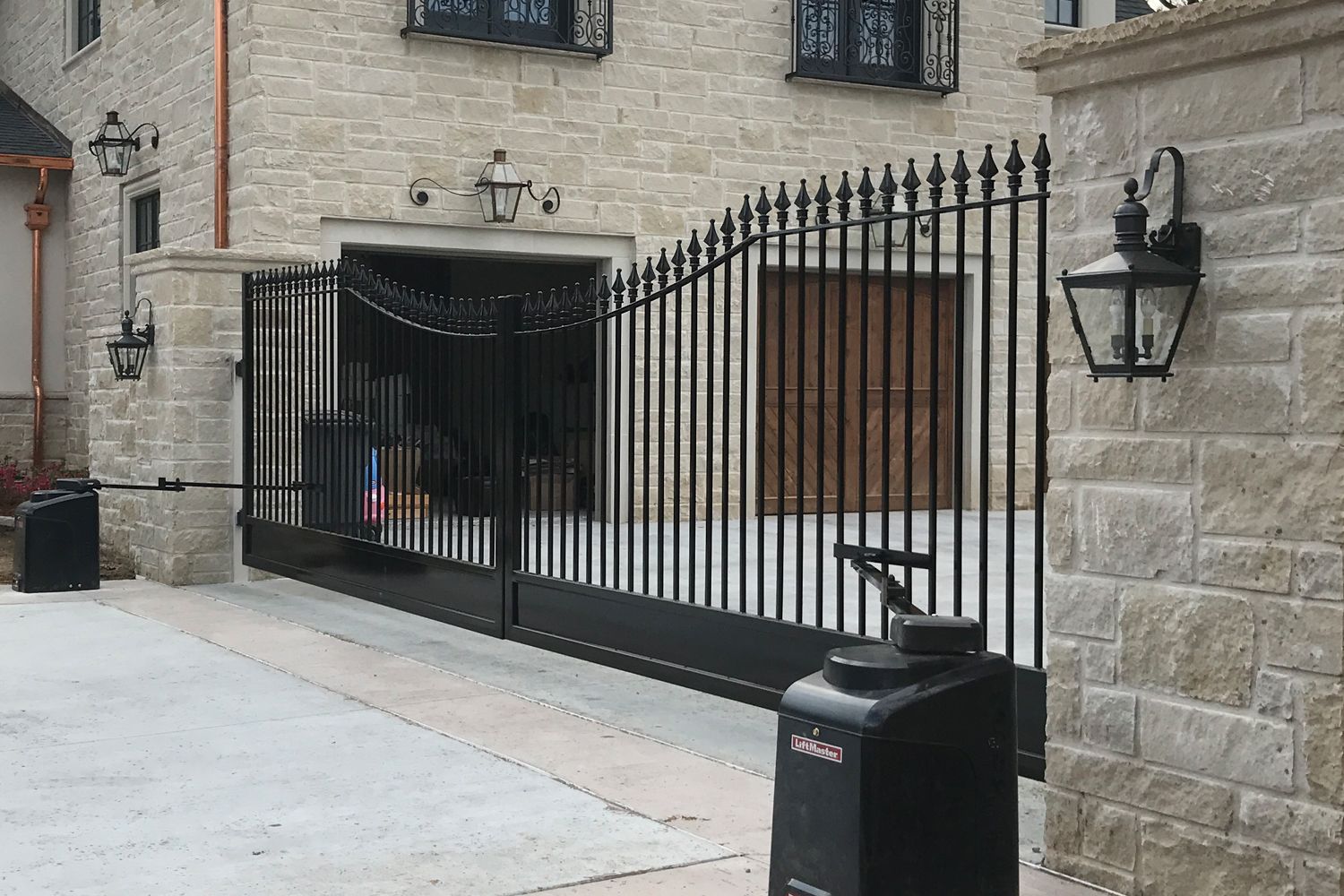 Black wrought iron driveway gate opening into a stone building with garage.