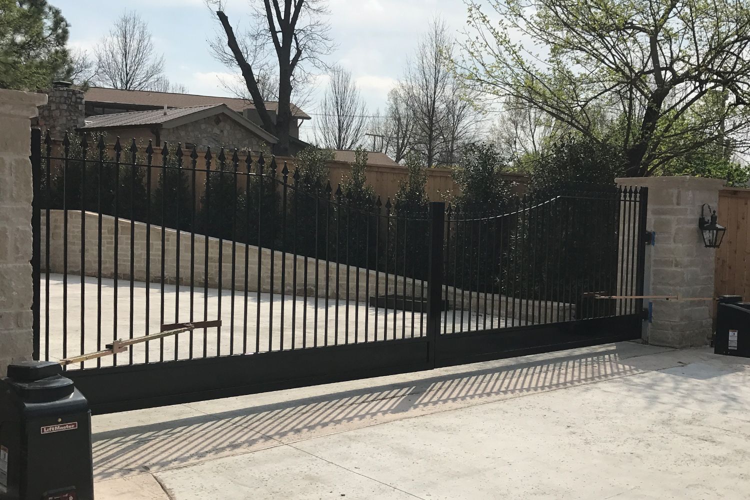 Black wrought iron gate with vertical bars, stone pillars, and a house in the background.