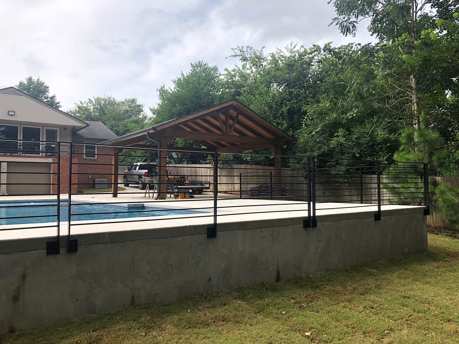 Black metal fence around a concrete pool deck. A wooden gazebo sits at the pool's edge, trees in the background.