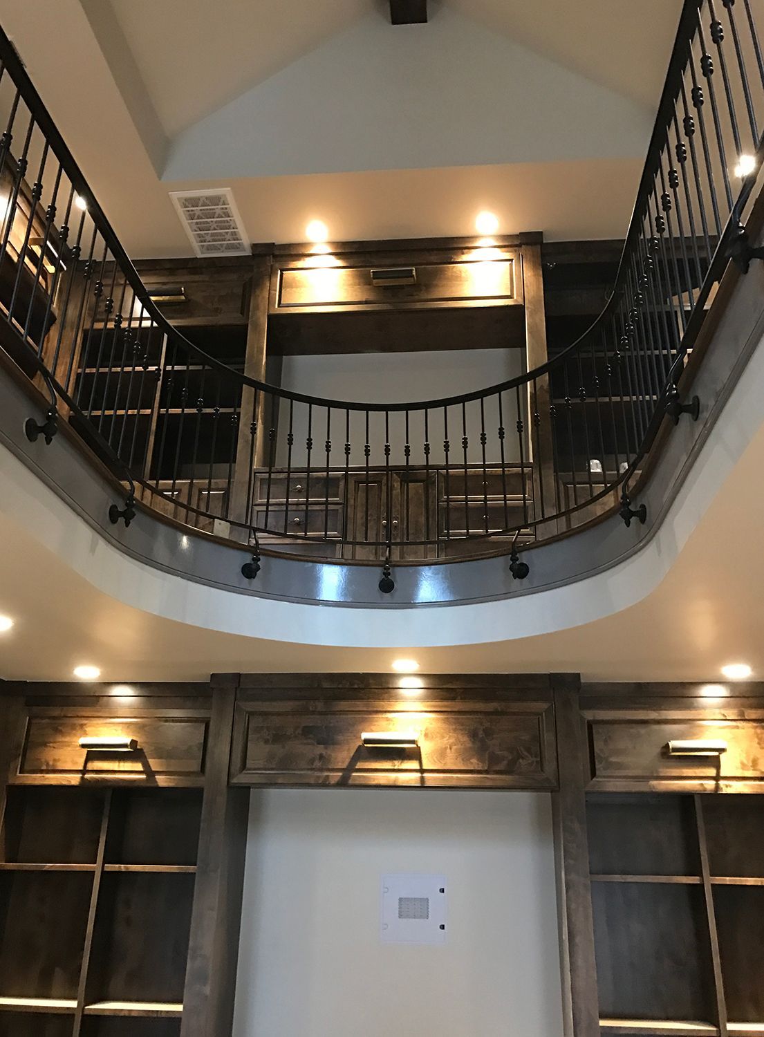 Wooden bookshelves spanning two levels with a wrought iron railing, lit by recessed lights.