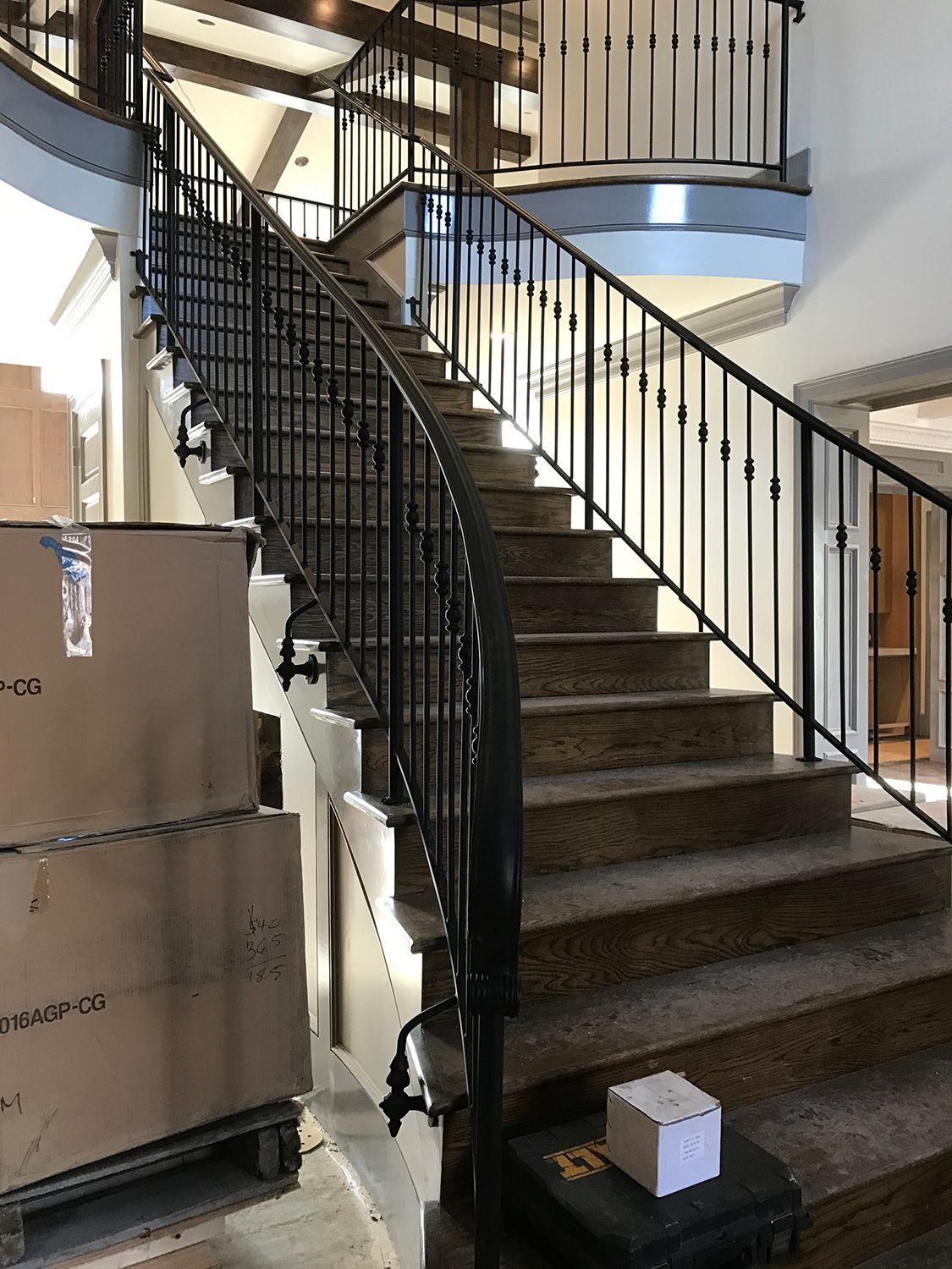 A curved staircase with black metal railing. Boxes on pallets are to the left, and a bright room in the background.