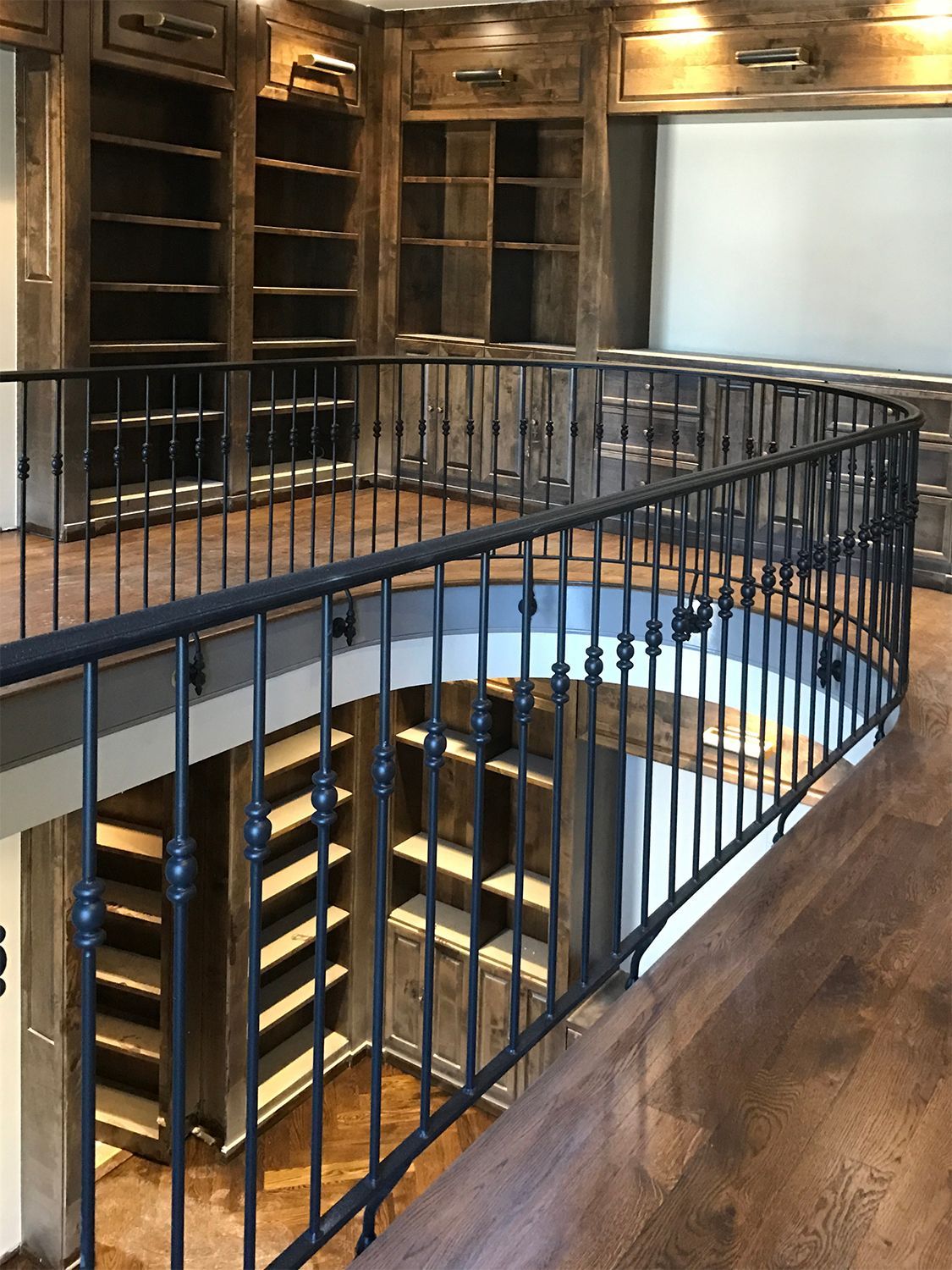 Two-story library with a wrought-iron railing. Wooden shelves are filled. Brown, tan, and gray tones dominate.