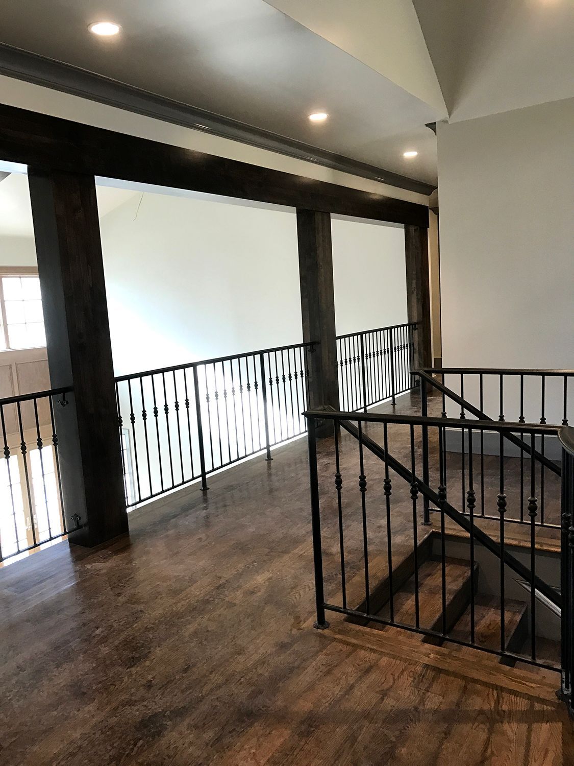 Interior shot of a second-floor hallway with dark wood flooring, metal railings, and a view into a room with large beams.
