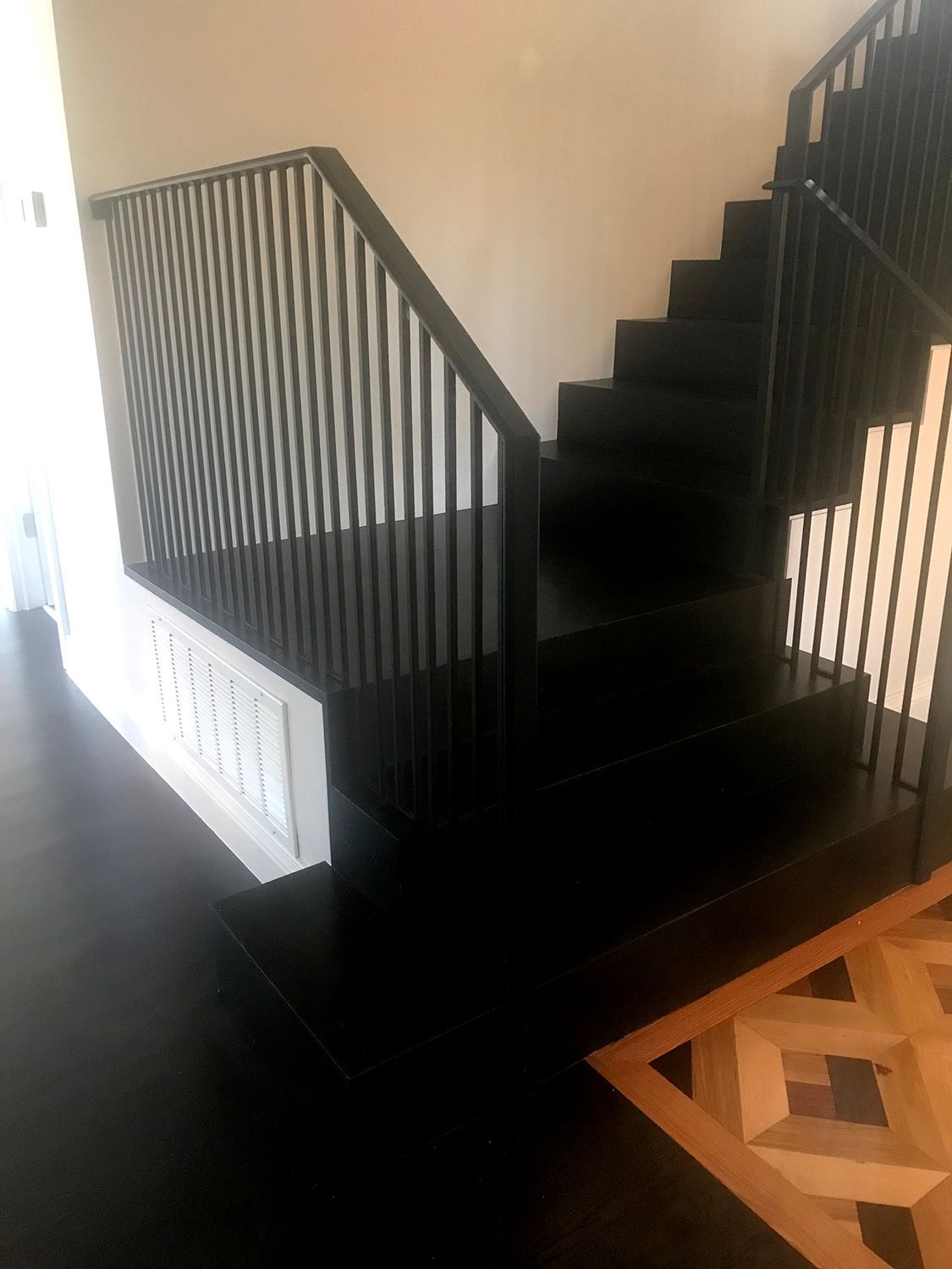 Black staircase with vertical black railings, white wall, and parquet flooring.