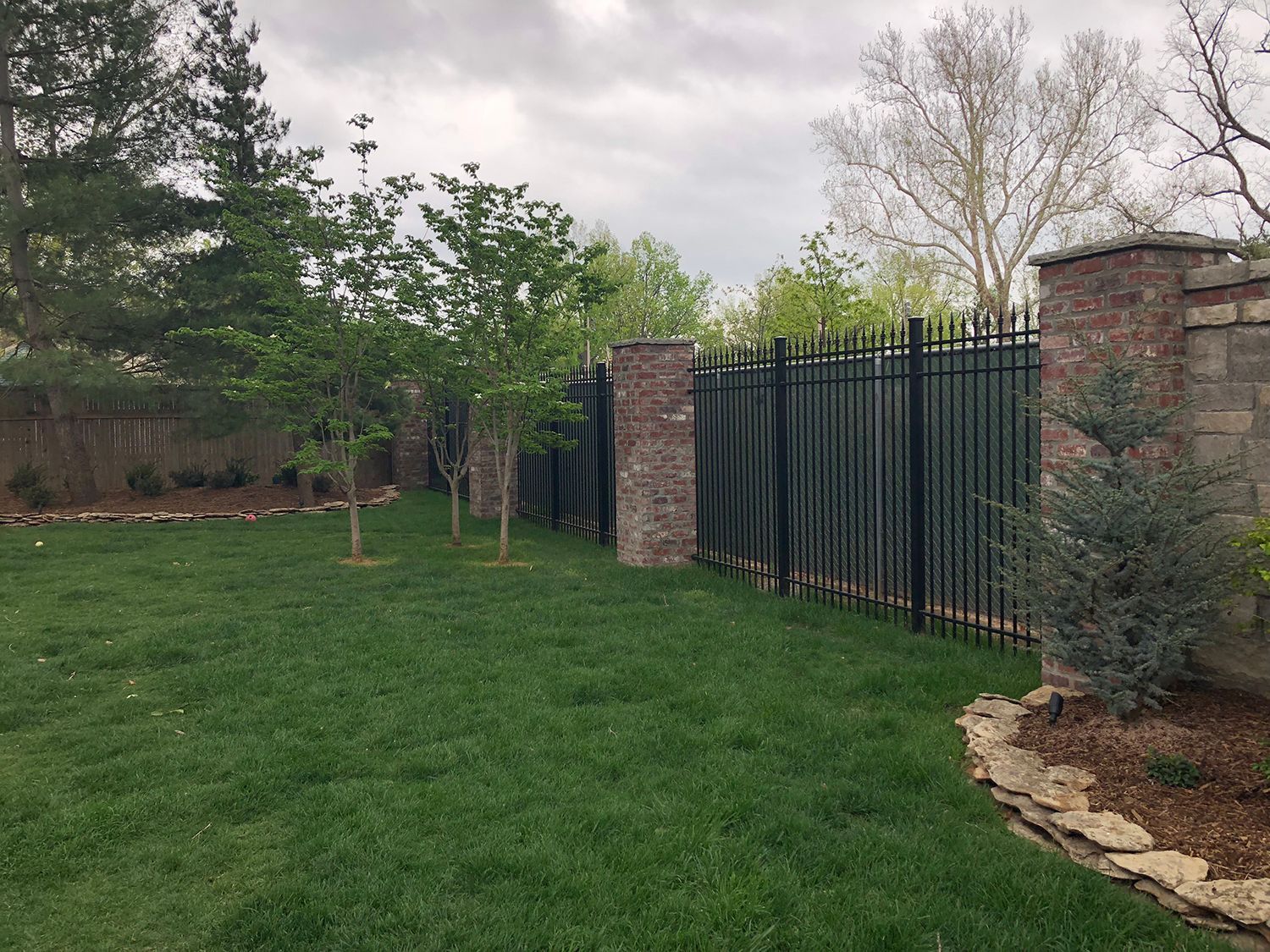Green lawn with a black metal fence, brick pillars, and trees under an overcast sky.