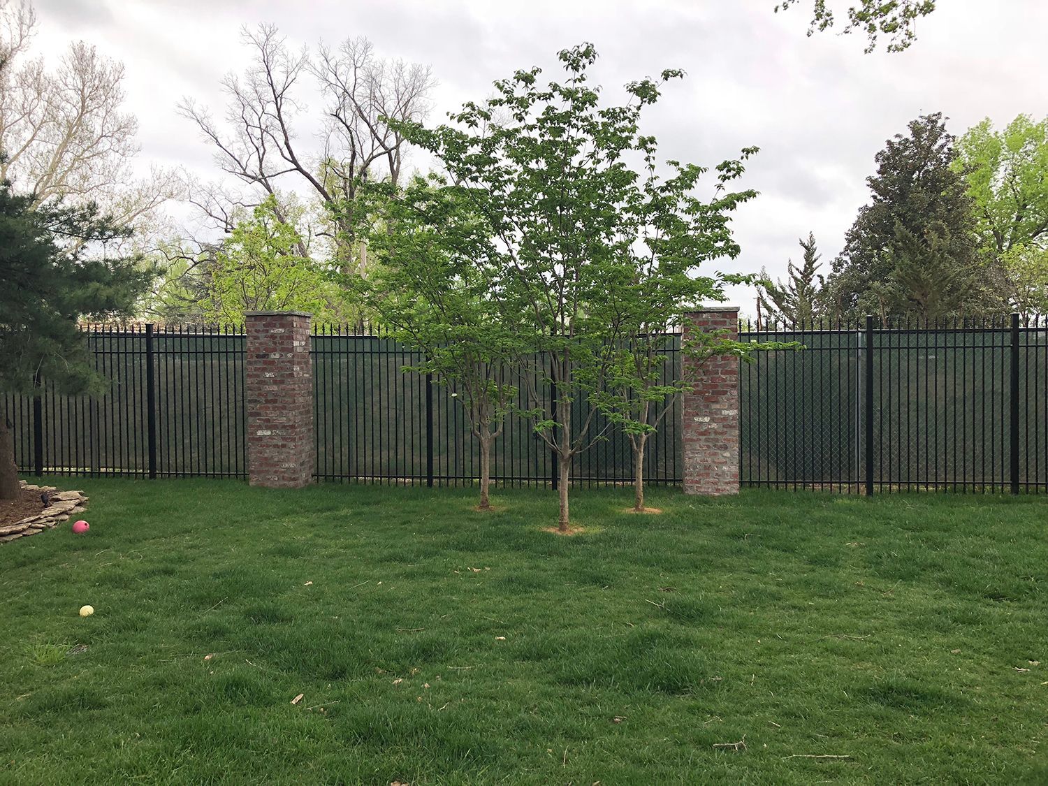 Black metal fence with brick pillars, trees, and green lawn in a backyard setting.