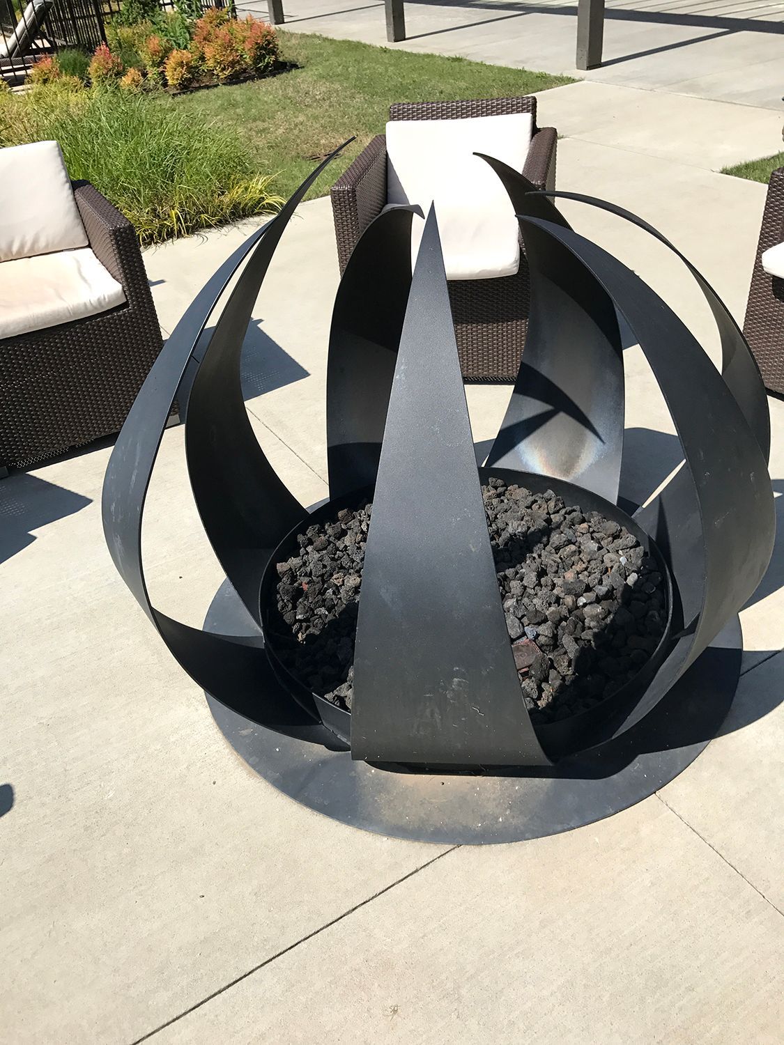 Black, orb-shaped fire pit on a concrete patio, surrounded by wicker chairs with white cushions.