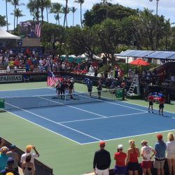 Tennis match with players, crowd, and American flag on a blue court under a sunny sky.