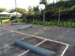 Rolls of chain link fencing on a weathered basketball court. A hoop, green vegetation, and a house are in the background.